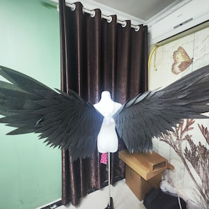 May include: Large, dark grey costume wings with a feathered appearance are displayed on a white mannequin. The wings are spread wide, showcasing their intricate design and texture. The background includes a green wall and brown curtains.