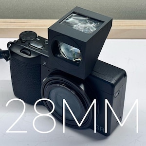 Puede incluir: Una cámara digital negra con una lente de 28 mm y un visor adjunto. La cámara tiene un agarre texturizado y una tapa de lente. El visor es rectangular e inclinado hacia arriba. El texto "28MM" se muestra en blanco.