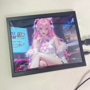 Może przedstawiać: Czarny monitor z ramką wyświetlający cyfrową ilustrację postaci anime z różowymi włosami, która nosi słuchawki. Postać siedzi na krześle z panoramą miasta w tle. Monitor wyświetla również pulpit z paskiem zadań i ikonami.