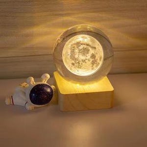 Op de afbeelding: Een glazen maanglobe met een wit maanontwerp erin, die op een houten voet staat met warm wit licht. De voet heeft een klein lampje dat de maanglobe verlicht.
