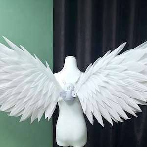 Pode incluir: Um par de asas grandes, brancas e pontiagudas, semelhantes a penas, presas a um arnês central num torso de manequim branco. As asas apresentam múltiplas camadas de penas brancas rígidas, alongadas e afiadas, estendendo-se numa ampla envergadura.