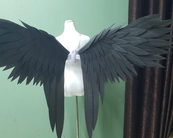 May include: A pair of large, black, feathered-style wings mounted on a white mannequin torso. The wings feature layered, pointed, dark fabric segments that mimic the appearance of bird feathers, extending outward from a central white harness.
