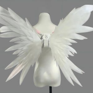Puede incluir: Alas de ángel blancas sobre un maniquí. Las alas están hechas de plumas translúcidas en capas, creando una apariencia delicada y etérea. Se adjuntan a un arnés con correas ajustables.