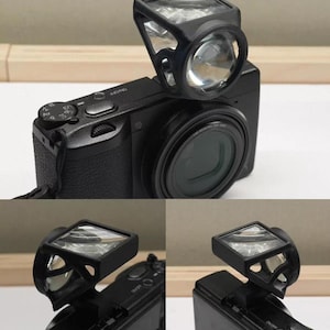 Puede incluir: Una cámara digital negra con un visor óptico montado. El accesorio tiene un marco negro con un elemento de lente de vidrio transparente en ángulo, colocado directamente sobre el cuerpo de la cámara para permitir una visión óptica directa.