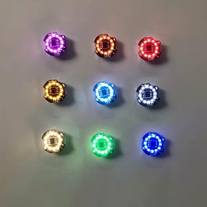 Puede incluir: Nueve módulos electrónicos circulares dispuestos en una cuadrícula, cada uno con un microchip negro central rodeado por un anillo de pequeños LED iluminados. Las luces brillan en varios colores, incluyendo morado, naranja, rojo, amarillo, azul claro, blanco, blanco cálido, verde y azul oscuro.