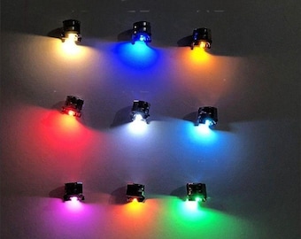 May include: Nine small, black LED lights emitting various colors. The lights are arranged in a grid pattern, with each light shining a different color, including yellow, blue, orange, red, white, purple, and green. The lights are on a dark gray background.