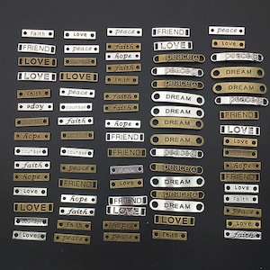 May include: A collection of 36 silver and gold-toned metal tags with various words and phrases, including "love", "peace", "faith", "hope", "friend", "courage", "dream", and "joy".