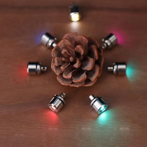 Op de afbeelding: Een bruine dennenappel wordt omringd door kleine, zilverkleurige LED-lampjes die verschillende kleuren uitstralen, waaronder rood, groen, blauw en geel. De lampjes zijn op een houten oppervlak geplaatst en creëren een decoratieve weergave.