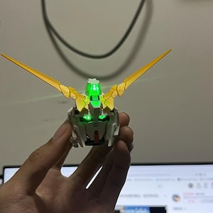 Peut inclure: Tête de modèle Gundam blanche et dorée avec des yeux et une crête verts brillants. Le modèle a des V-fins jaunes s'étendant de la tête. La tête est tenue dans une main, avec un écran d'ordinateur visible en arrière-plan.