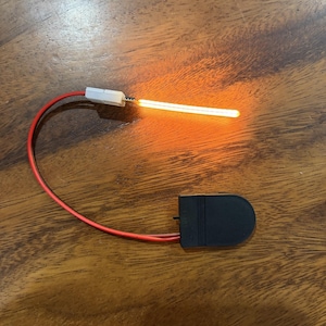 May include: An illuminated, orange-glowing light strip with a red wire and a black battery holder. The light strip is straight and emits a warm, bright light. The components are set against a wooden surface.