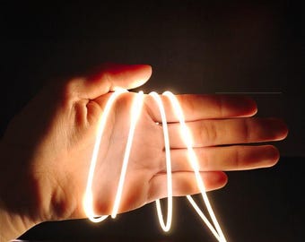 May include: A hand holding a glowing, white, flexible light cord. The cord is coiled and illuminated against a dark background, creating a bright, ethereal effect. The light appears to be a soft, warm white.