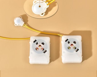 May include: Two white, square-shaped electronic components with circular patterns and small black rectangles. A yellow wire connects them to a smaller, white component with a bear-shaped design. The background is a solid tan color.
