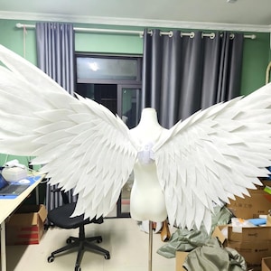 Pode incluir: Grandes asas de fantasia brancas com um design de penas em camadas. As asas são exibidas em um manequim, mostrando seu tamanho e forma. Ideais para cosplay ou uso teatral.