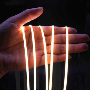 Könnte beinhalten: Nahaufnahme einer Hand, die mehrere leuchtende, dünne, weiße LED-Lichtstreifen hält. Die Lichter strahlen ein warmes, gelb-oranges Leuchten vor einem dunklen Hintergrund aus. Das Bild zeigt die Flexibilität und Helligkeit der LED-Streifen.