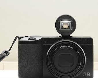 May include: A black digital camera with a textured grip and a lens featuring the text LOS XSLM-PRO DMC NANO. The camera has a circular external flash unit attached to the top and the letters GR printed in the bottom right corner of the body.