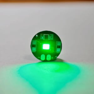 May include: A circular green printed circuit board featuring a central bright white light-emitting diode. The board includes two rectangular contact pads on the sides and two oval-shaped connection points at the bottom marked with plus and minus signs.