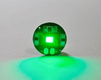 May include: A circular green printed circuit board featuring a central bright white light-emitting diode. The board includes two rectangular contact pads on the sides and two oval-shaped connection points at the bottom marked with plus and minus signs.