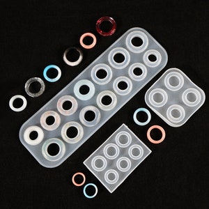 May include: Two silicone molds for making resin rings. The larger mold has 15 round cavities, while the smaller mold has 6 round cavities. There are also several colorful resin rings scattered around the molds.