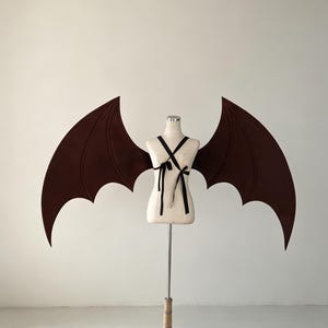 May include: A pair of large, dark brown bat wings attached to a beige mannequin torso. The wings feature a pointed, scalloped edge design with visible structural lines. The mannequin is secured with black ribbon straps that cross over the chest.