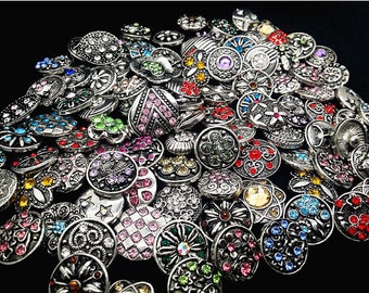 May include: A large collection of silver-toned snap buttons with various colorful rhinestone embellishments. The buttons feature a variety of designs, including floral, geometric, and abstract patterns.