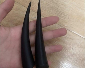 May include: A pair of matte black, curved, tapered horn-shaped objects. Each piece features a smooth, solid surface with a pointed tip and a flat, circular base. The items are elongated and possess a distinct, symmetrical, and sleek design.