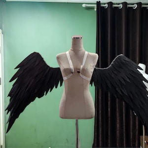 May include: A pair of large, black, feathered-style wings attached to a beige mannequin torso. The wings are secured with white elastic straps that cross over the chest, featuring a central dark circular accent piece on the harness.