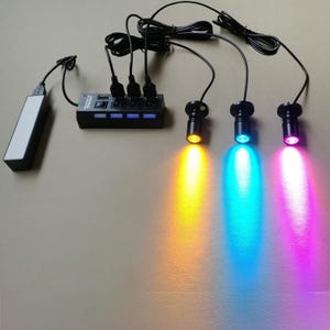 May include: A black USB hub with four ports and three LED spotlights emitting yellow, blue, and pink light. The hub is connected to a power bank via a USB cable, and the spotlights are connected to the hub.