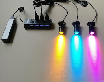 May include: A black USB hub with four ports and three LED spotlights emitting yellow, blue, and pink light. The hub is connected to a power bank via a USB cable, and the spotlights are connected to the hub.