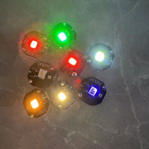 Puede incluir: Una colección de pequeñas luces LED redondas en varios colores, incluyendo rojo, verde, blanco, amarillo y azul. Cada luz está montada en una pequeña placa de circuito negro con el texto "JX2533". Las luces están dispuestas sobre una superficie gris.