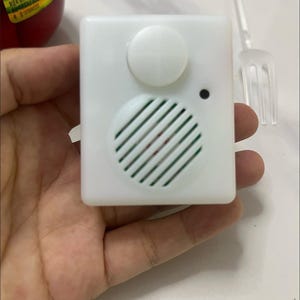 May include: A small, white, square sound module featuring a speaker grill, a button, and a black dot. The module is held in a hand, with a clear plastic fork in the background. The product label is partially visible.