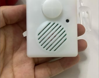 May include: A small, white, square-shaped sound module with a speaker grill, a button, and a small black dot. The module is held in a hand, with a clear plastic fork in the background. The product label is partially visible.