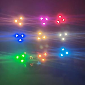 May include: A grid of nine light clusters arranged in three rows and three columns. Each cluster consists of three small, square, glowing light sources emitting vibrant colours including orange, pink, red, blue, yellow, white, green, and amber.