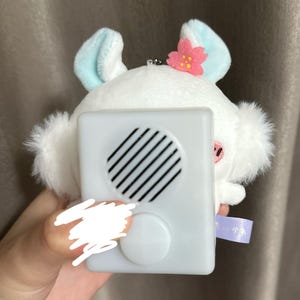 以下が含まれることがあります： 白いうさぎのぬいぐるみ。水色の耳とピンクの花の飾りがある。白いスピーカーボックスが取り付けられており、丸いグリルとボタンが付いている。日本語のテキストが書かれた小さな紫色のタグが見える。