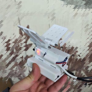 Può includere: Un dispositivo bianco stampato in 3D con più pannelli incernierati e una superficie perforata. È dotato di una luce rossa e fili scoperti. Un cavo bianco e nero è collegato al dispositivo.