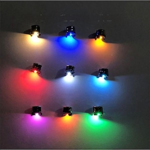Op de afbeelding: Negen kleine, zwarte LED-lampjes die verschillende kleuren uitstralen. De lampjes zijn gerangschikt in een rasterpatroon, waarbij elk lampje een andere kleur schijnt, waaronder geel, blauw, oranje, rood, wit, paars en groen. Op een donkergrijze achtergrond.