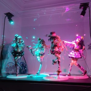 Op de afbeelding: Vier anime-stijl beeldjes tentoongesteld in een glazen vitrine, verlicht door blauwe en roze lichten. De beeldjes staan op witte cirkelvormige bases, elk met unieke outfits en poses. De vitrine staat op een donkere ondergrond.