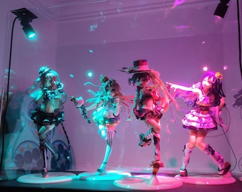 May include: Four anime-style figurines displayed in a glass case, illuminated by blue and pink lights. The figurines are of young women in colorful, detailed outfits, each posed on a circular base. The case is on a dark surface.