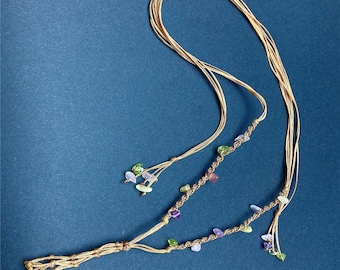 May include: A handmade macrame necklace with a tan cord and colorful gemstone accents. The necklace features strands of cord with small, colorful stones in shades of green, purple, and clear. The necklace is displayed on a dark blue background.