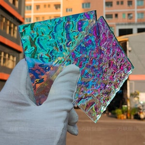 May include: Two iridescent glass panels with a textured surface are held by a gloved hand. The panels display a vibrant array of colors, including teal, purple, and gold, reflecting light in a dynamic pattern. The background shows a building.