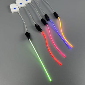 Pode incluir: Várias mechas de luz de fibra ótica com várias cores, incluindo verde, vermelho, rosa, azul e amarelo. Cada mecha tem um conector preto e uma base de plástico branco com um botão azul. As mechas estão dispostas diagonalmente sobre um fundo cinza.