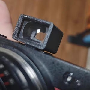 Puede incluir: Un visor rectangular negro fijado a una cámara. El visor tiene una abertura cuadrada y está hecho de un material texturizado. El cuerpo de la cámara también es negro, con detalles plateados.