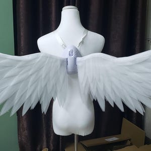 May include: A pair of large, white, articulated wings attached to a central white harness worn on a mannequin torso. The wings feature multiple layers of pointed, feather-like segments that extend horizontally, creating a wide, expansive wingspan.