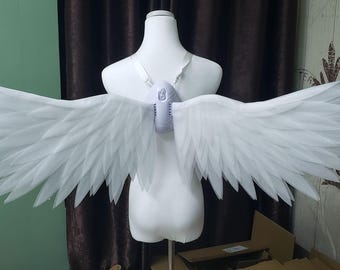 May include: A pair of large, white, articulated wings attached to a central white harness worn on a mannequin torso. The wings feature multiple layers of pointed, feather-like segments that extend horizontally, creating a wide, expansive wingspan.