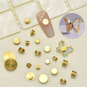May include: A collection of gold-colored nail art supplies, including round studs and butterfly-shaped embellishments. The studs vary in size and shape, with some having a cylindrical design. One nail is decorated with a gold stud and pearls.