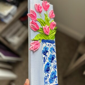 May include: A textured painting of pink tulips in a blue and white vase. The vase has a floral pattern. The background is a light blue color. The painting is on a rectangular canvas.