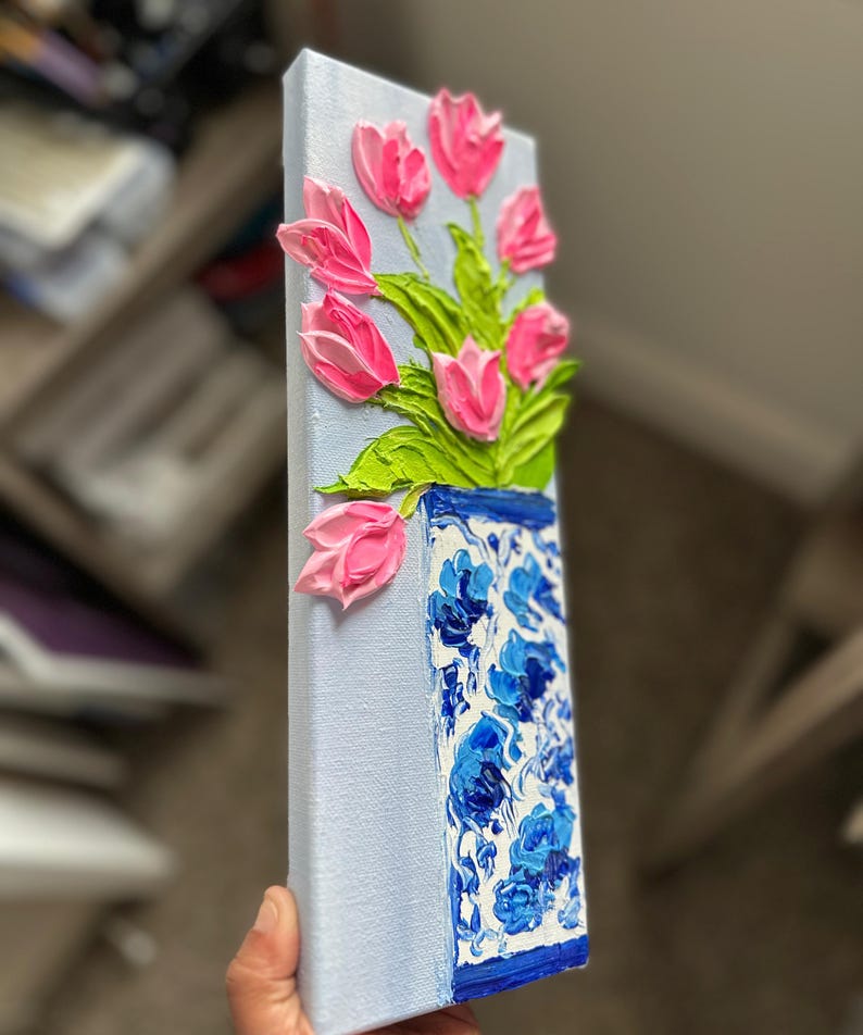 May include: A textured painting of pink tulips in a blue and white vase. The vase has a floral pattern. The background is a light blue color. The painting is on a rectangular canvas.