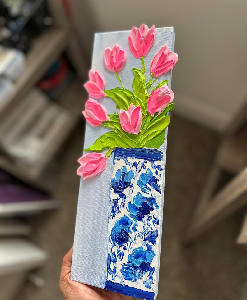 May include: A rectangular canvas painting featuring a vase of pink tulips. The vase is blue and white with a floral pattern. The tulips have green leaves and are painted with a textured, raised effect, creating a 3D appearance.