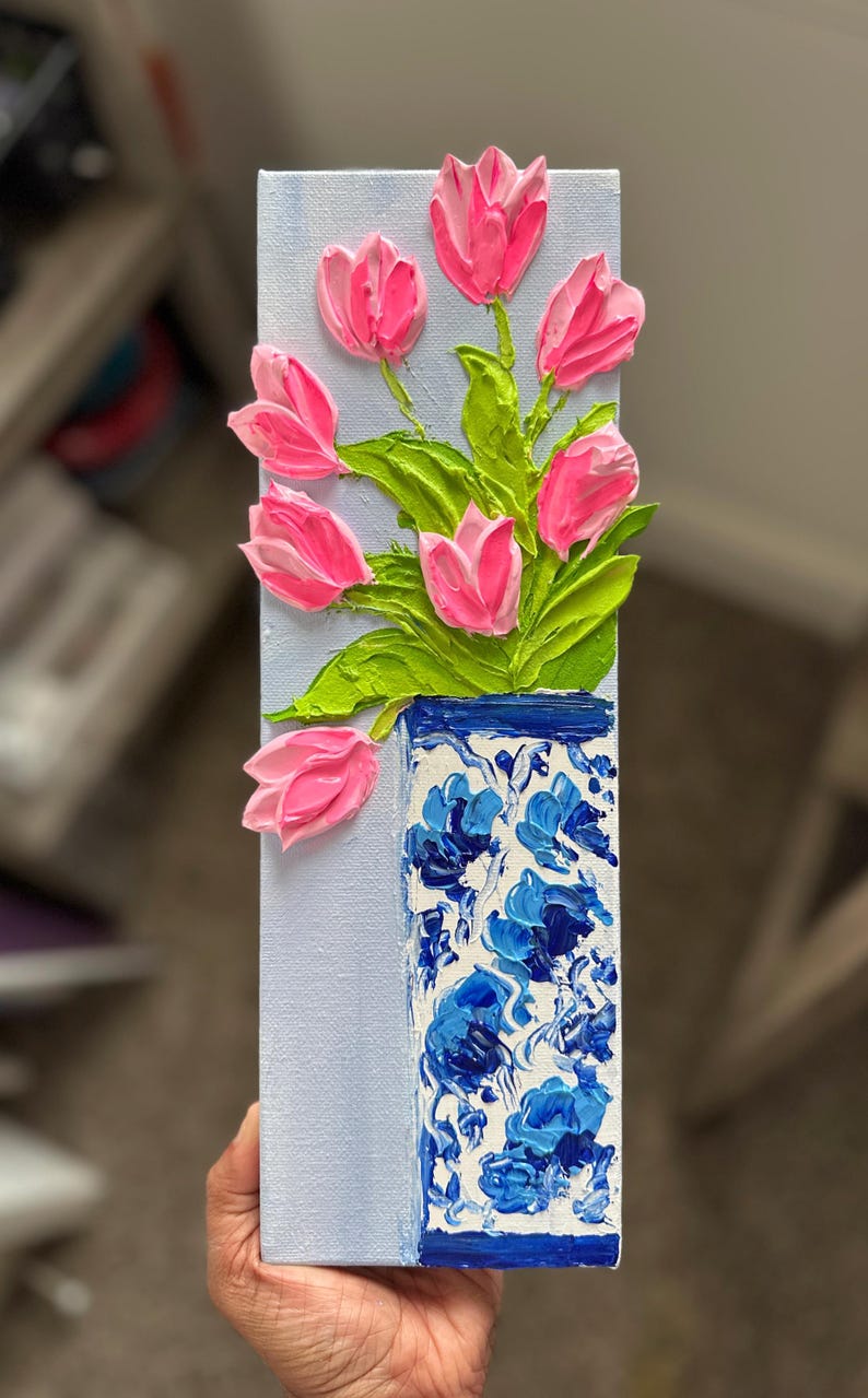 May include: A vertical painting of pink tulips in a blue and white vase. The tulips are textured and stand out from the light blue background. The vase has a floral pattern.