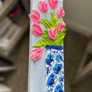 May include: A vertical canvas painting featuring a bouquet of pink tulips in a blue and white vase. The tulips have textured petals and green leaves. The vase has a floral pattern and is set against a light blue background.