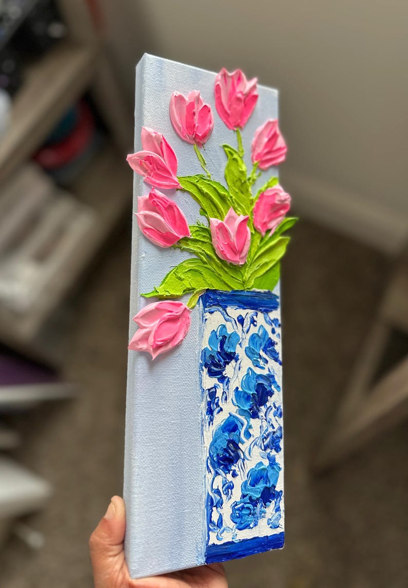 May include: A vertical canvas painting featuring a bouquet of pink tulips in a blue and white vase. The tulips have textured petals and green leaves. The vase has a floral pattern and is set against a light blue background.